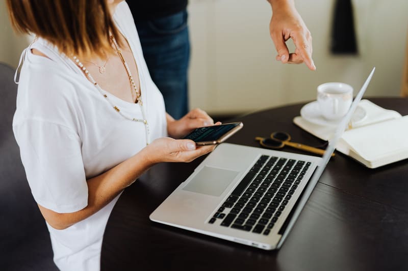 Dueña de un negocio revisa en el móvil su landing page mientras el diseñador le señala cambios en el portátil