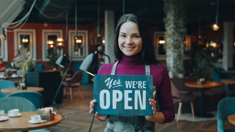 Propietaria de una cafetería junto al cartel de abierto